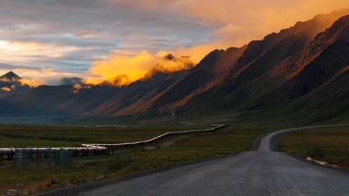 A winding road leads through a valley with mountains, under a dramatic sunset sky with clouds and warm colors.