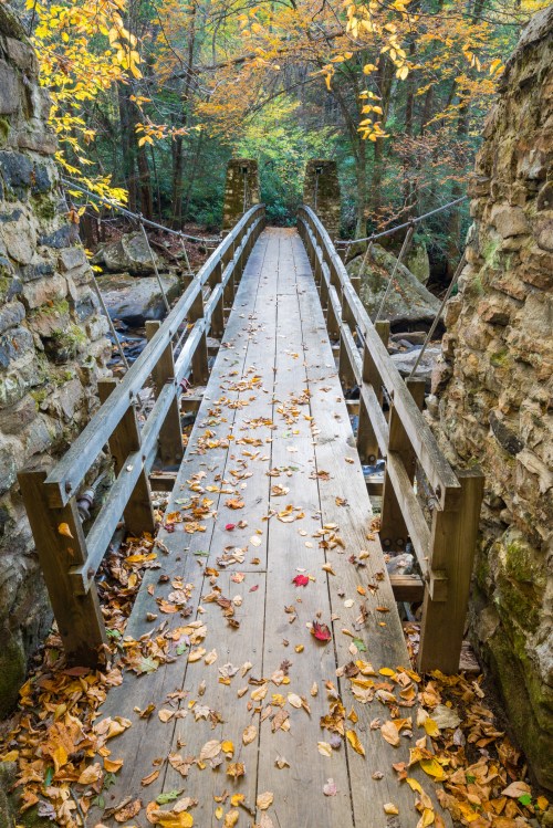 A wooden bridge covered in autumn leaves, surrounded by stone walls and vibrant fall foliage in a forested area.