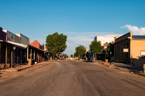 A quiet, empty street in a small town with historic buildings and trees under a clear blue sky.