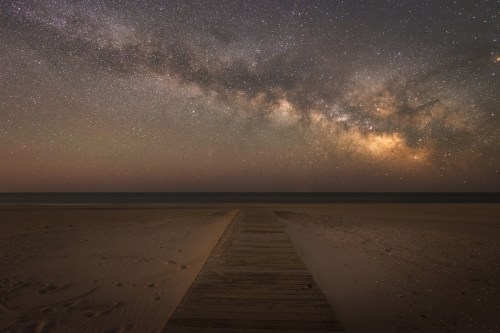 A wooden path leads to a serene beach under a starry night sky filled with the Milky Way.