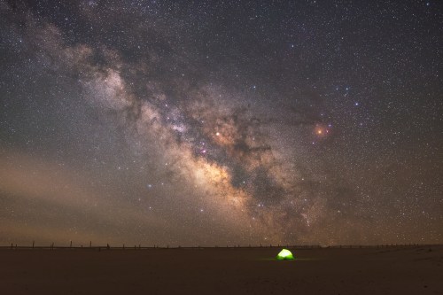 A green tent under a starry sky, showcasing the Milky Way and a vast desert landscape.