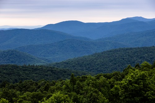 A panoramic view of rolling green mountains under a cloudy sky, showcasing layers of hills and dense forest.