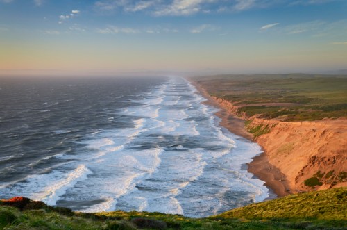 A scenic view of a coastline with rolling waves, sandy beaches, and lush green cliffs under a colorful sky.