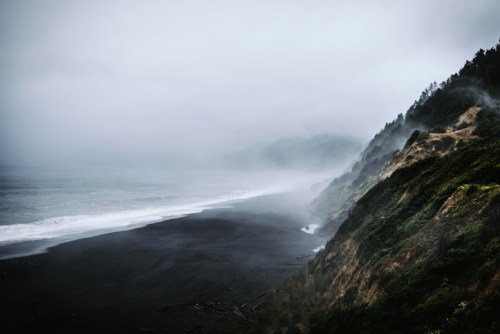 A misty coastal scene with dark sand, waves, and a steep, green hillside under a cloudy sky.