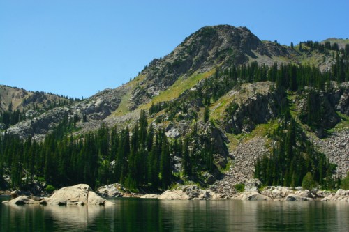 A serene mountain landscape with lush green trees and rocky terrain reflected in calm water under a clear blue sky.