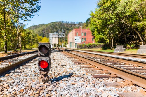 A railway signal stands beside train tracks, with a red light illuminated, surrounded by trees and a distant building.