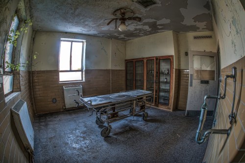 Abandoned room with a gurney, peeling paint, and a window, evoking a sense of neglect and decay.