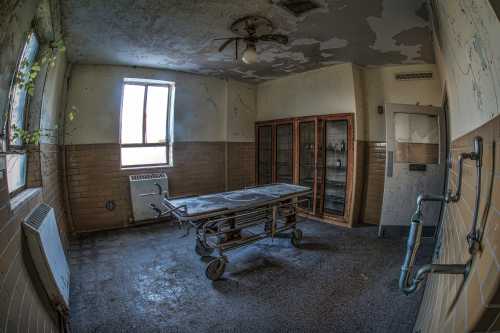 Abandoned room with a gurney, peeling paint, and a window, evoking a sense of neglect and decay.