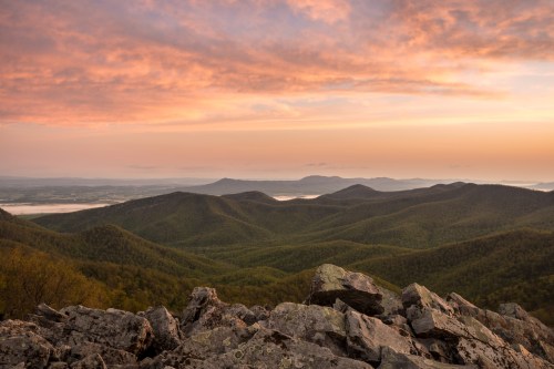 A scenic view of rolling green mountains under a colorful sunset sky, with rocky foreground and distant hills.