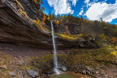A scenic waterfall cascading down rocky cliffs, surrounded by autumn foliage and a clear blue sky.