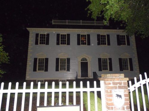 A historic two-story house at night, featuring white walls, black shutters, and a white picket fence.