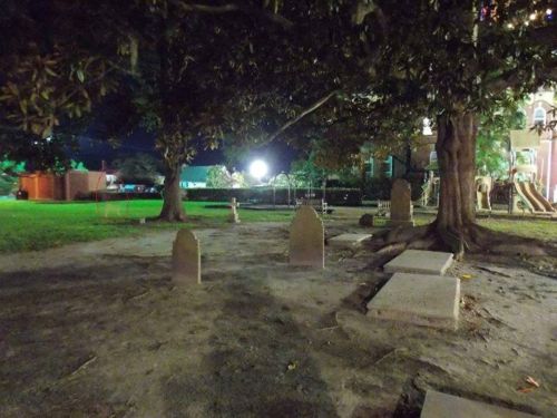 A dimly lit graveyard at night, featuring several gravestones surrounded by trees and a grassy area.