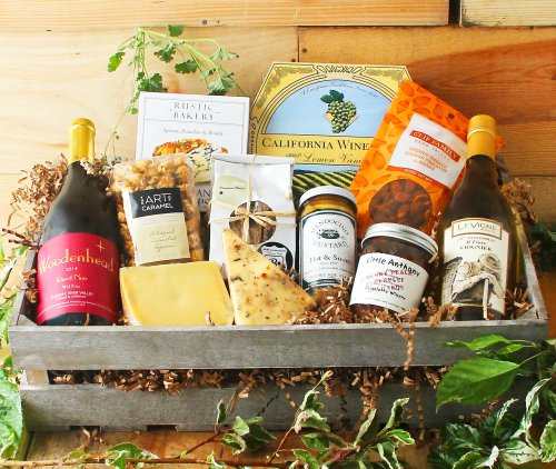A wooden basket filled with wine, cheese, snacks, and gourmet treats, surrounded by greenery.