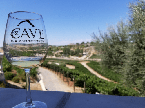 A glass of white wine with "The Cave Oak Mountain Winery" logo, overlooking a vineyard under a clear blue sky.
