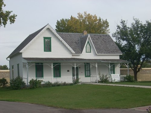 A white, two-story house with green trim, a porch, and a well-maintained lawn surrounded by trees.