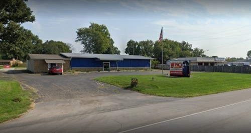 A blue building with a flag, surrounded by grass and trees, with a sign in front on a paved area.