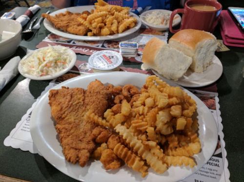 A plate of fried fish, crinkle-cut fries, and popcorn shrimp, with coleslaw and rolls on the side.
