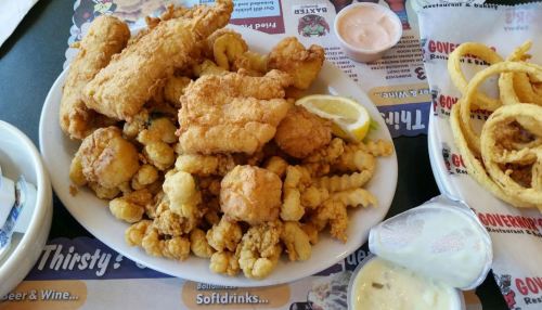 A plate of fried seafood including fish, shrimp, and calamari, served with lemon and sides of onion rings and dipping sauce.