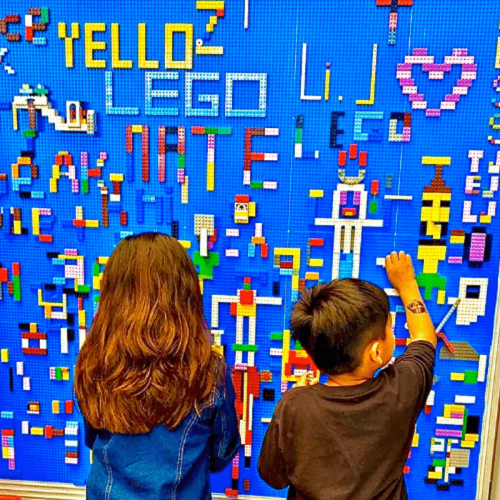 Two children create art on a colorful LEGO wall, surrounded by various LEGO structures and designs.