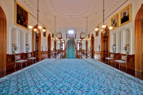 A grand hallway with a staircase, ornate decor, and portraits on the walls, featuring a patterned carpet.