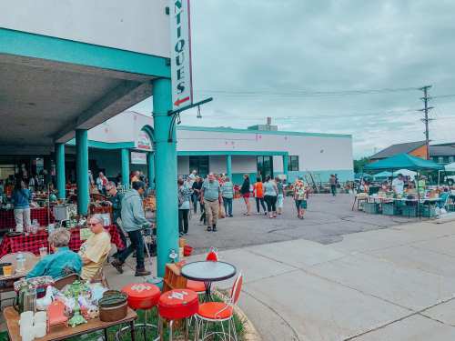 A bustling outdoor market with people browsing antiques and goods, surrounded by colorful tents and tables.