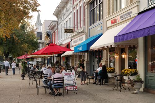 Outdoor dining scene with people at tables, colorful awnings, and shops lining a charming street in autumn.