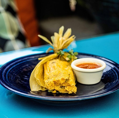 A tamale wrapped in corn husks on a blue plate, accompanied by a small bowl of red sauce.