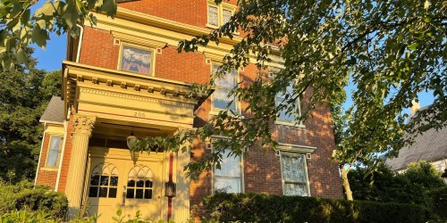 A large brick house with yellow trim, surrounded by greenery, under a clear blue sky.