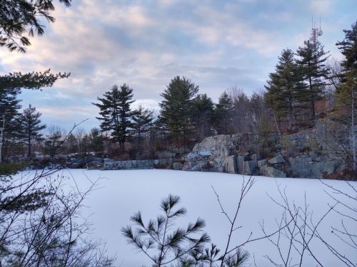 A serene winter scene featuring a frozen lake surrounded by rocky cliffs and evergreen trees under a cloudy sky.