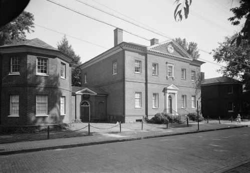 Historic brick building with a symmetrical facade, surrounded by trees and a paved street, in a quiet neighborhood.