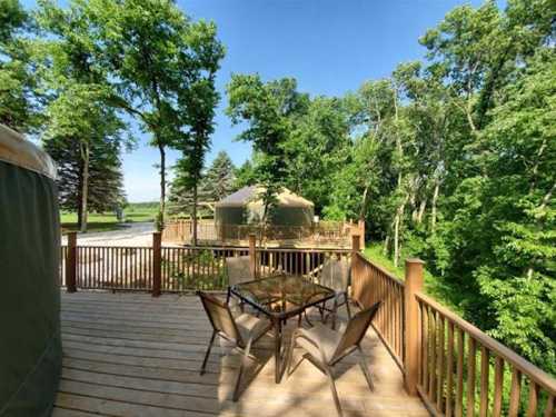 A wooden deck with a glass table and chairs, surrounded by lush green trees and a yurt in the background.