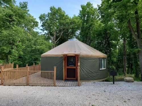 A yurt with a wooden deck surrounded by trees, set in a gravel area.