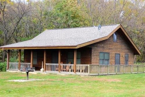 A rustic wooden cabin with a porch, surrounded by trees and grass, featuring a grill in the foreground.