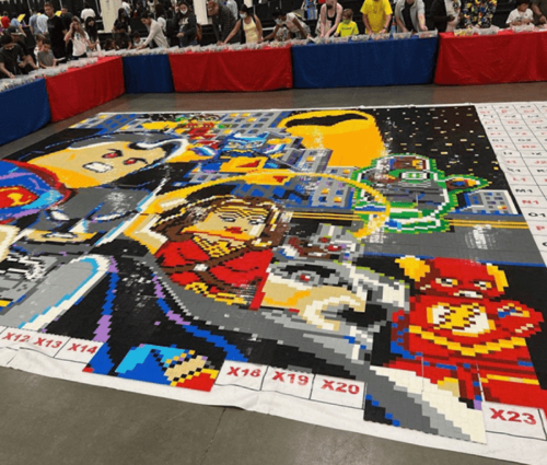 A large, colorful LEGO mosaic featuring superhero characters in a cityscape, displayed at a crowded event.