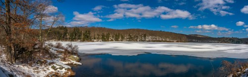 A serene winter landscape featuring a frozen lake surrounded by trees under a bright blue sky with fluffy clouds.