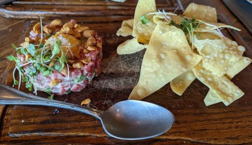A wooden platter with beef tartare topped with nuts and an egg yolk, alongside crispy tortilla chips.
