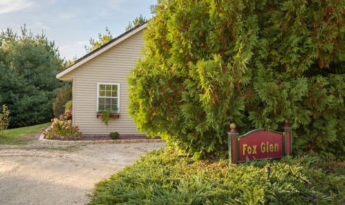 A cozy house with a sign reading "Fox Glen," surrounded by lush greenery and a gravel path.
