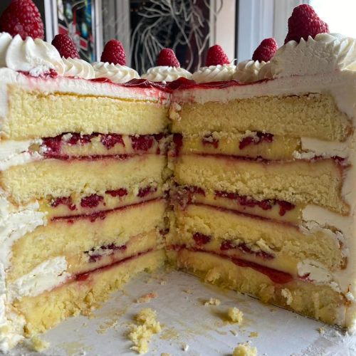A layered cake with yellow sponge, raspberry filling, and whipped cream, topped with fresh raspberries.