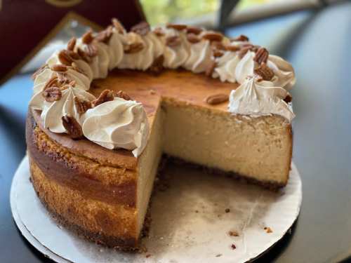 A creamy cheesecake topped with whipped cream and pecans, with a slice cut out, sitting on a white plate.