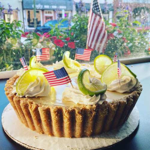 A decorated pie with lime slices and small American flags, set against a window with flowers and an American flag outside.