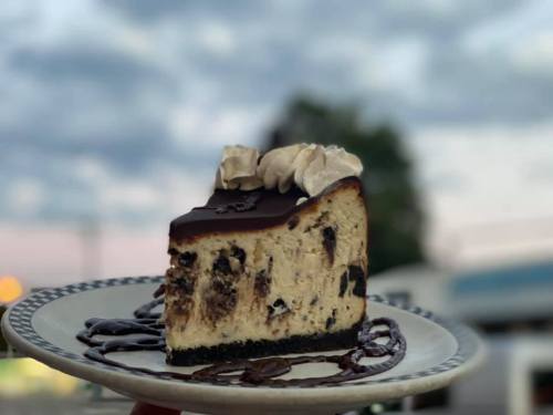A slice of chocolate cheesecake topped with whipped cream, served on a plate against a blurred outdoor background.
