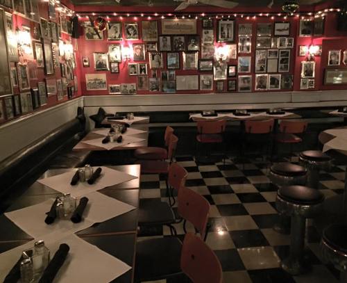 Cozy restaurant interior with red walls, framed photos, checkered floor, and tables set with napkins and condiments.