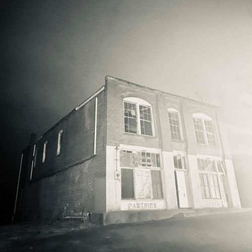 Abandoned brick building with large windows, partially obscured by fog, featuring a faded "Pastries" sign.