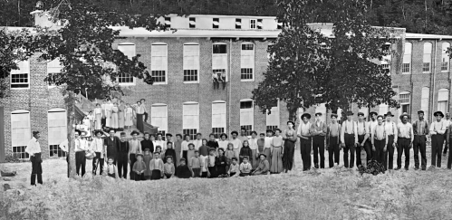 A large group of people, including children and adults, poses in front of a brick building with many windows.