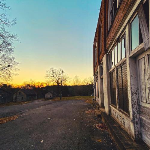 An abandoned building at sunset, with trees and distant structures in a serene landscape.