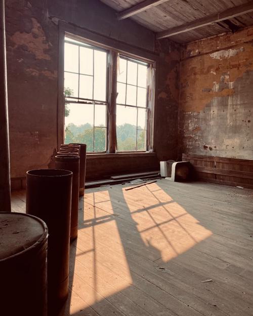 Sunlight streams through large windows, casting shadows on a rustic, empty room with wooden floors and peeling walls.