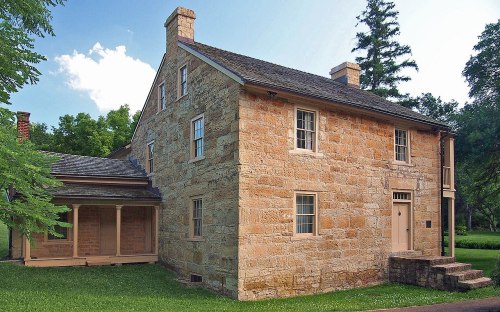 Historic stone house with a sloped roof, surrounded by greenery and trees under a blue sky.