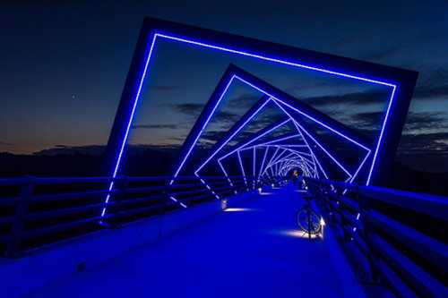 A lit bridge at night, featuring blue neon lights in a geometric pattern, with a bike parked on the side.