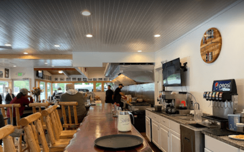 Interior of a casual restaurant with a wooden bar, kitchen area, and patrons seated at the counter.