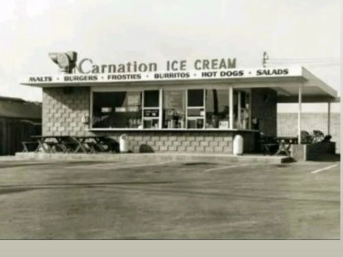 A vintage ice cream shop with a sign for malts, burgers, and more, featuring outdoor seating and a classic design.